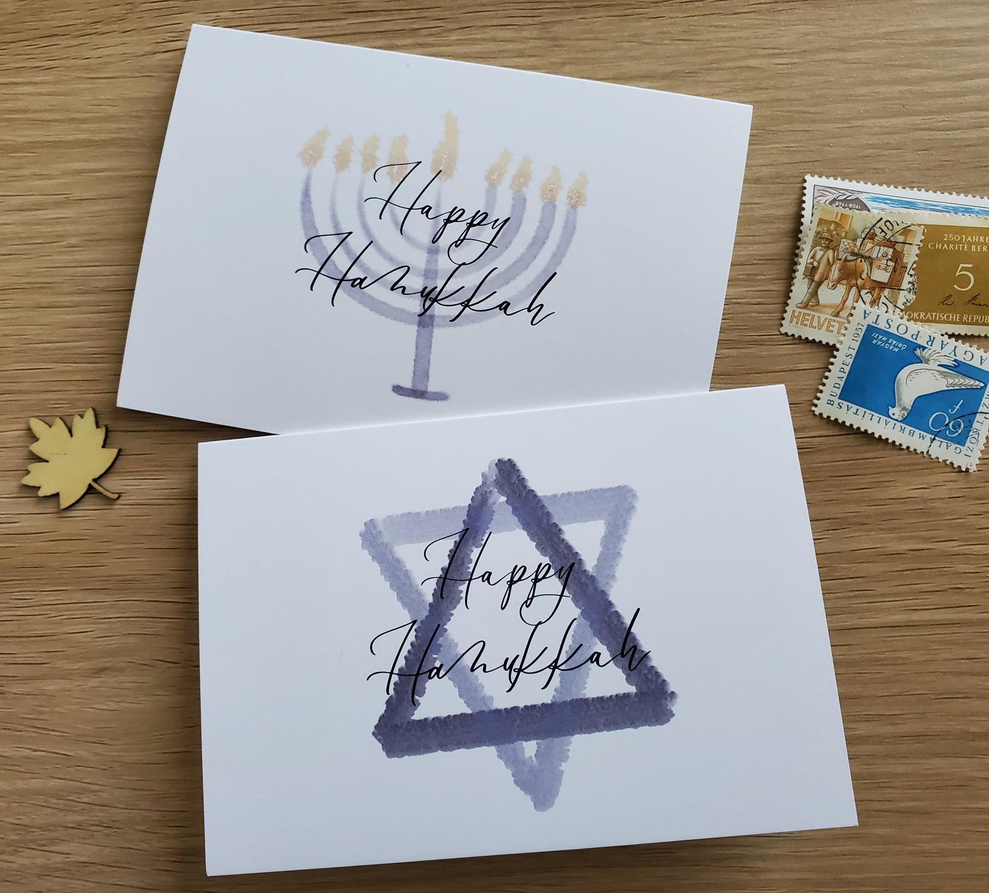 photo of watercolor notecards in landscape orientation, sitting flat on a wood table . card features a watercolor illustration of either a menorah or star of David and message "Happy hanukkah"
