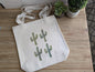 Tote bag with a cactus watercolor design featuring 4 cacti on a wooden surface.